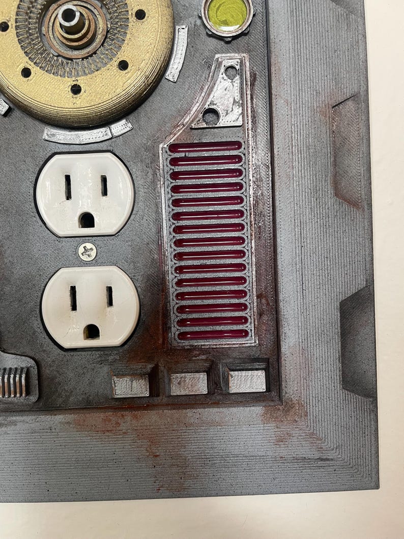 May include: A close-up of a gray, industrial-style wall plate with two white electrical outlets. The plate features metallic accents, including a circular gear-like element and a section with red bars. The overall design has a steampunk aesthetic.