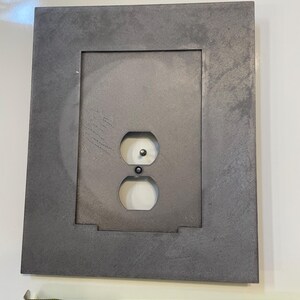 May include: A gray metal electrical outlet cover with a rectangular frame. The cover has two outlet holes and a grounding hole. A measuring tape shows the cover is approximately 6 inches wide.