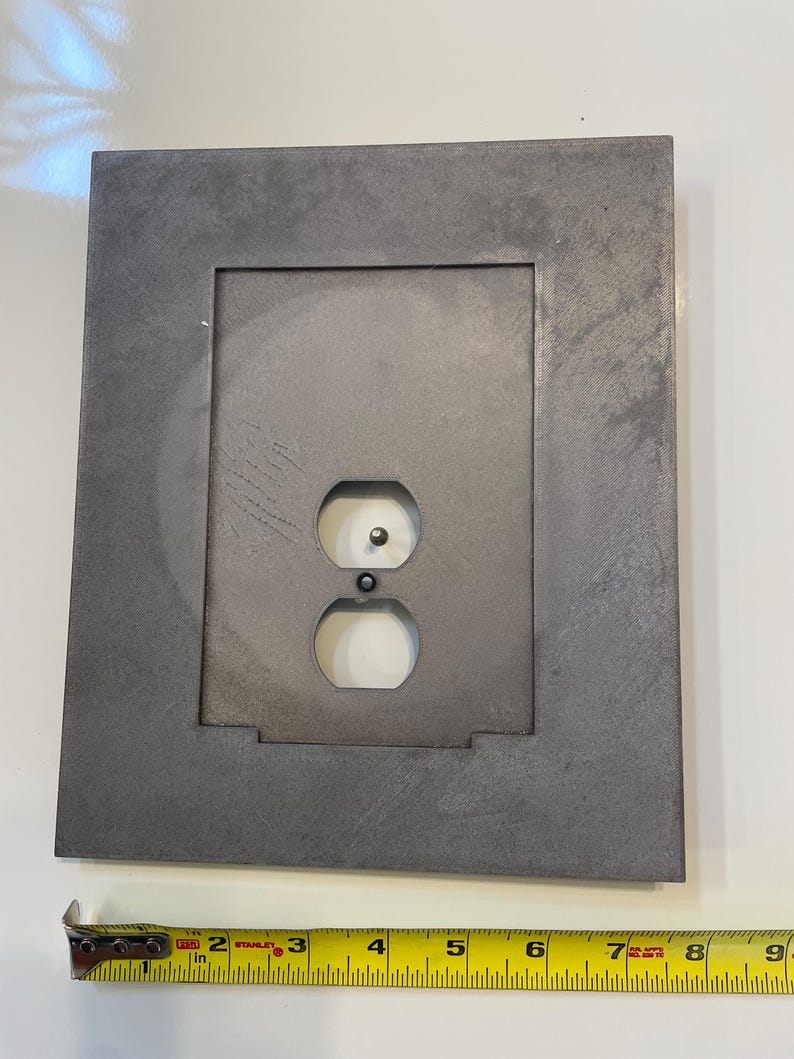 May include: A gray metal electrical outlet cover with a rectangular frame. The cover has two outlet holes and a grounding hole. A measuring tape shows the cover is approximately 6 inches wide.