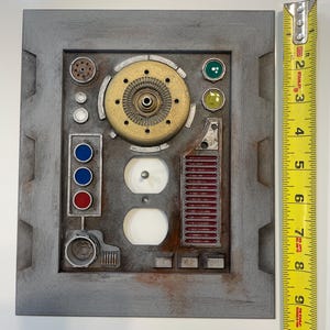 May include: A gray, rectangular panel with various mechanical components, including a brass-colored circular piece, red and blue buttons, and two white outlets. A measuring tape shows the panel is approximately 10 inches in length.