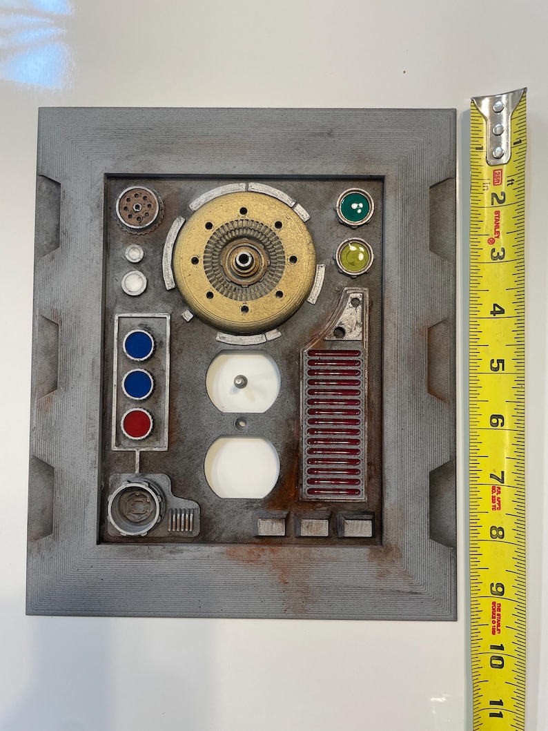 May include: A gray, rectangular panel with various mechanical components, including a brass-colored circular piece, red and blue buttons, and two white outlets. A measuring tape shows the panel is approximately 10 inches in length.