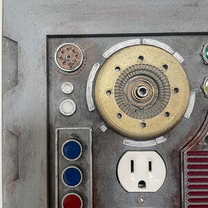 May include: A detailed, industrial-style wall art piece featuring a metallic gray frame. The artwork incorporates various mechanical components, including a brass gear, white electrical outlets, and colored button lights. The design has a steampunk aesthetic.