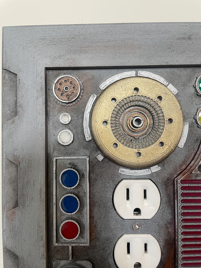 May include: A detailed, industrial-style wall art piece featuring a metallic gray frame. The artwork incorporates various mechanical components, including a brass gear, white electrical outlets, and colored button lights. The design has a steampunk aesthetic.