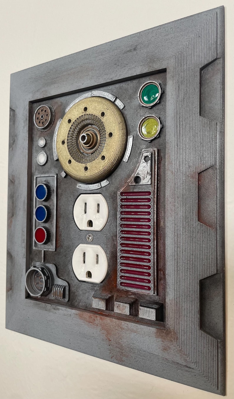 May include: A gray, rectangular wall art piece with a steampunk aesthetic. It features a variety of mechanical-looking components, including a brass-colored circular element, two white electrical outlets, and red and green accents. The frame has a textured, weathered appearance.