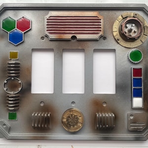 May include: A metallic gray light switch cover with a weathered, industrial design. It features three switch openings, along with various colored geometric shapes, circular elements, and a series of rectangular and circular details. The color palette includes shades of gray, brown, red, green, blue, and gold.
