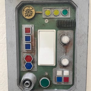 May include: A weathered, gray control panel with a green interior. It features a white light switch, various colored buttons, and a circular gold emblem. The panel has a vintage, industrial aesthetic.