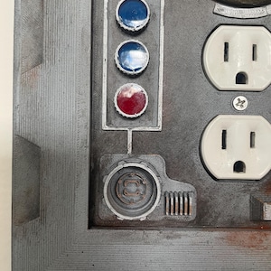 May include: A close-up of a gray, industrial-style panel with various components. It features three circular lights, two blue and one red, and two white electrical outlets. The panel has a textured surface and a metallic appearance.
