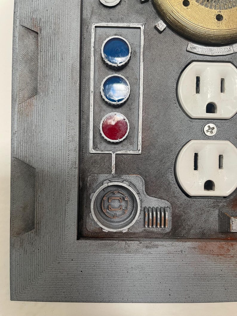 May include: A close-up of a gray, industrial-style panel with various components. It features three circular lights, two blue and one red, and two white electrical outlets. The panel has a textured surface and a metallic appearance.