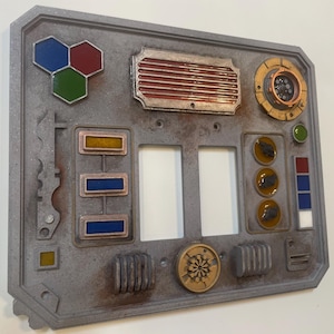 May include: A gray, weathered-looking light switch cover with various colored geometric shapes and panels. Features a circular dial, three toggle switches, and a series of colored rectangles. The cover has a retro, industrial design.