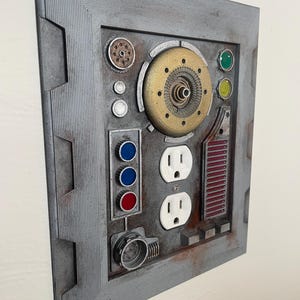 May include: A gray, rectangular wall panel with a variety of embedded components. It features two electrical outlets, colored buttons, and a circular gold-colored mechanism. The panel has a textured, industrial design with a weathered appearance.