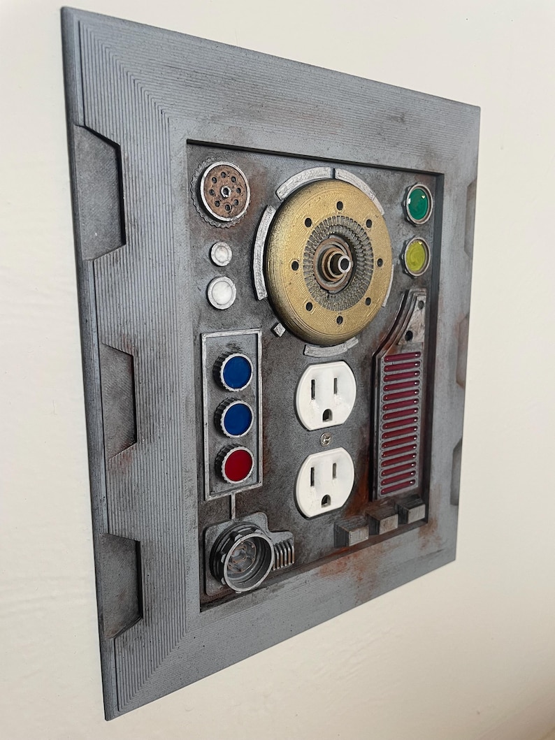 May include: A gray, rectangular wall panel with a variety of embedded components. It features two electrical outlets, colored buttons, and a circular gold-colored mechanism. The panel has a textured, industrial design with a weathered appearance.