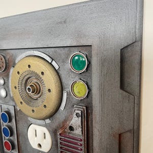 May include: A detailed, industrial-style wall art piece. It features a gray, textured frame and a panel with various components, including electrical outlets, buttons, and a circular, brass-colored element. Green and yellow circular accents add color.