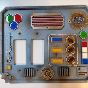 May include: A rectangular gray control panel featuring a metallic finish with various raised elements including a hexagonal green, red, and blue emblem, a red-slatted vent, circular dials, rectangular buttons, and two large empty rectangular cutouts.