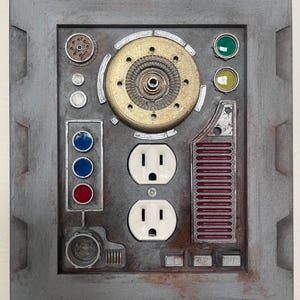 May include: A gray, industrial-style wall plate with various mechanical components. It features two electrical outlets, multiple colored indicator lights, and a central circular design with a gold-colored center. The plate has a weathered, metallic appearance.