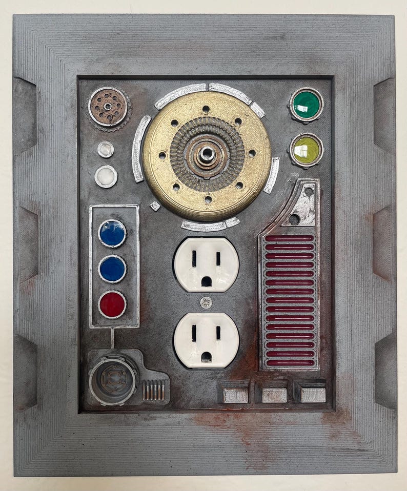 May include: A gray, industrial-style wall plate with various mechanical components. It features two electrical outlets, multiple colored indicator lights, and a central circular design with a gold-colored center. The plate has a weathered, metallic appearance.