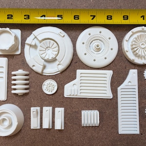 May include: A collection of off-white 3D-printed model parts, including circular components with intricate designs, rectangular pieces, and smaller gear-like elements. A yellow tape measure is placed above the parts, showing measurements in inches.