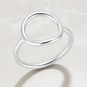 May include: A silver ring with a modern design. The ring features a circular top element connected to a band. The ring is made of a shiny, reflective metal, and is set against a neutral background.