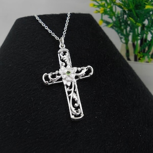 Op de afbeelding: Een zilveren kruis-hangerketting met een ingewikkeld bloemenontwerp. Het kruis heeft een kleine groene edelsteen in het midden van een bloem. De ketting wordt gepresenteerd op een zwart oppervlak.
