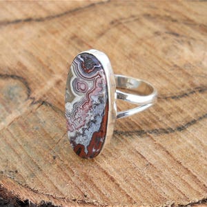 May include: A silver ring with an oval stone displaying a distinctive pattern of red, white, grey, and brown swirls. The ring has a split shank design, set against a wooden background.