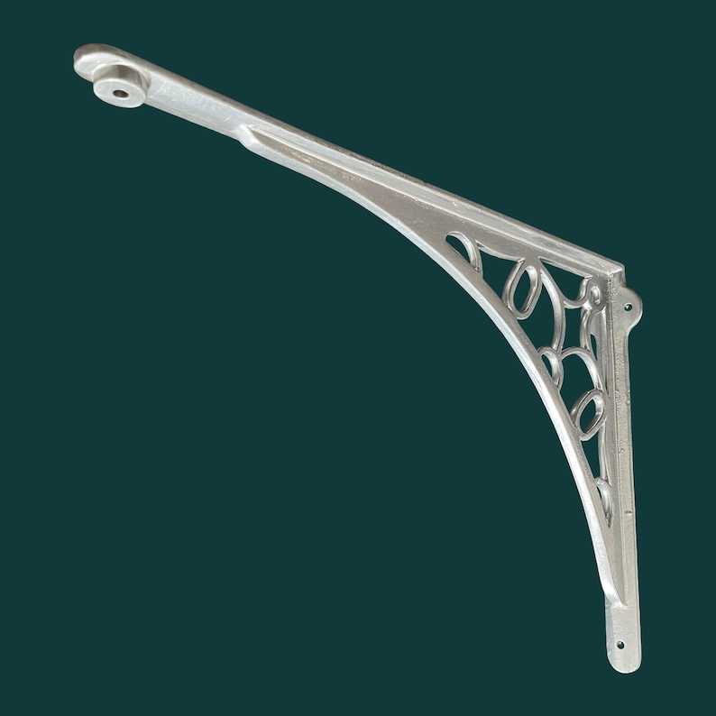 K&ouml;nnte beinhalten: Ein silberfarbener, dekorativer Regaltr&auml;ger mit einem bogenf&ouml;rmigen Design. Der Tr&auml;ger hat einen l&auml;nglichen Arm mit einem kreisf&ouml;rmigen Loch am Ende und eine vertikale Montageplatte. Der gebogene Abschnitt hat offene, wirbelnde Muster.