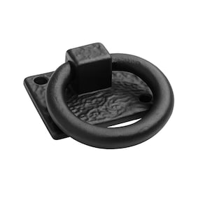 Może przedstawiać: Czarny uchwyt pierścieniowy z kwadratową podstawą i teksturowanym wykończeniem. Pierścień jest okrągły i przymocowany do kwadratowej podstawy z dwoma otworami montażowymi. Okucia są metalowe.