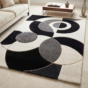 May include: A rectangular area rug with a geometric design. The rug features a pattern of black, white, and gray shapes, including circles and curved lines. The rug is placed on a light-colored floor, and a beige sofa is visible in the background.