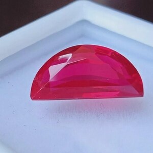 Peut inclure: Une pierre précieuse rouge vif, taillée en demi-lune et à facettes. La gemme est posée sur un fond blanc, mettant en valeur ses surfaces polies et ses reflets internes. La taille de la pierre permet à la lumière de jouer sur ses facettes, créant un éclat brillant.