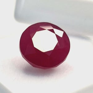 May include: A faceted, round ruby gemstone. The gem is a deep red color with multiple facets that reflect light. The ruby is set against a white background, highlighting its color and shape. This gemstone is suitable for jewelry.
