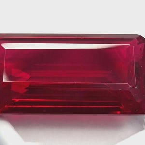 May include: A rectangular, faceted gemstone with a deep red hue. The gem has a clean, polished appearance with sharp edges and a reflective surface. The gemstone is set against a white background, highlighting its color and clarity. This is a jewelry component.