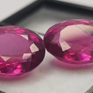 Puede incluir: Dos gemas facetadas de corte ovalado con un tono rosa intenso. Las gemas se exhiben sobre una superficie blanca dentro de un estuche negro. Las facetas capturan la luz, creando reflejos brillantes.