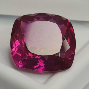 Puede incluir: Una gema facetada, de talla cojín, con un vibrante tono magenta. La gema tiene un gran centro de color claro y está colocada sobre un fondo blanco. El corte de la piedra crea múltiples facetas que reflejan la luz, realzando su brillo.