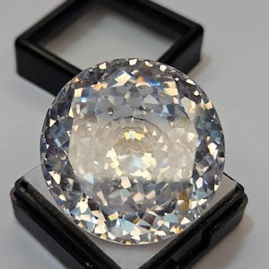 May include: A large, round, faceted gemstone with a brilliant, clear appearance. The gem is set in a black display case, showcasing its many reflective surfaces. The facets create a dazzling display of light and sparkle.