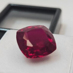 May include: A faceted, cushion-cut ruby gemstone. The gem is a deep, saturated red color, with multiple facets reflecting light. It is set against a white surface, with a black display case in the background. The gemstone is the focal point of the image.