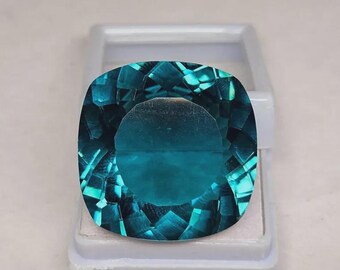 May include: A faceted, teal-colored gemstone with a cushion cut, displayed in a white plastic case. The gem's surface reflects light, showcasing its intricate internal structure and color variations. The overall shape is square with rounded corners.