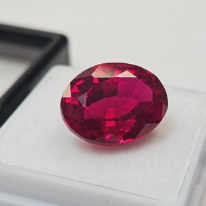 Puede incluir: Una gema de rubí tallada en forma ovalada y facetada. La gema es de un color rojo intenso y saturado, con múltiples facetas que reflejan la luz. El rubí está sobre un fondo blanco, con una caja negra parcialmente visible.