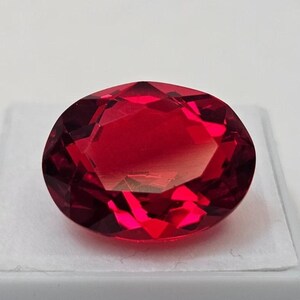 Peut inclure: Une pierre précieuse rouge foncé, taillée en forme ovale. La gemme est posée sur un fond blanc, mettant en valeur sa taille brillante et sa couleur vibrante. Les facettes de la pierre précieuse reflètent la lumière.