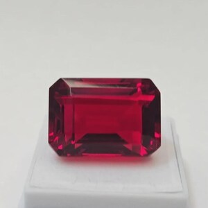 May include: A faceted, rectangular-cut, deep red gemstone. The gem is displayed on a white surface against a white background. The gemstone's facets reflect light, creating a brilliant appearance. The cut is an emerald cut.