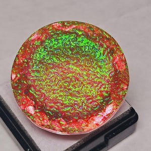 May include: A round, iridescent gemstone with a textured surface. The gem features a vibrant color palette of red, green, and hints of yellow. The stone is set on a black base, showcasing its unique color and pattern.
