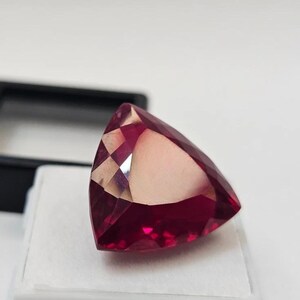 May include: A faceted, triangular-shaped gemstone with a deep red hue. The gem is displayed on a white surface, with a black display case in the background. The gem's cut creates multiple reflective surfaces.