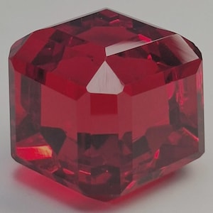Op de afbeelding: Een gefacetteerde, robijnrode edelsteen met een geometrische, kubusachtige vorm. De vele facetten van de edelsteen reflecteren het licht, waardoor een briljante weergave van kleur en glans ontstaat. De edelsteen is diep, rijk rood.