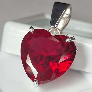 Puede incluir: Un colgante en forma de corazón con una gran gema facetada de color rojo intenso. La gema está sujeta por cuatro garras plateadas y tiene una argolla plateada para una cadena. El colgante está sobre un fondo blanco.