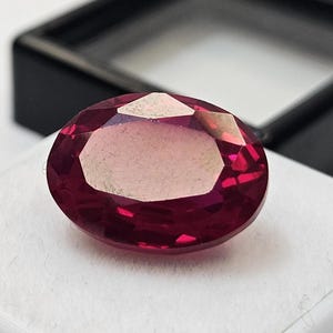 May include: A faceted, oval-cut ruby gemstone. The gem is a deep red colour with a lighter, almost pinkish centre. It sits on a white surface, with a black display case in the background.
