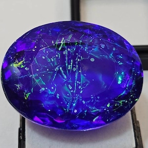 May include: An oval-shaped gemstone with a dark blue hue, displaying internal patterns of green and white. The polished surface reflects light, revealing intricate details. The gemstone is set against a dark backdrop, accentuating its vibrant colours and internal features.