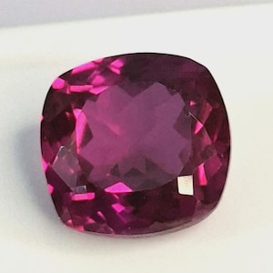 May include: A faceted, cushion-cut gemstone with a deep magenta color. The gem has a polished surface and reflects light, showcasing its internal structure. The shape is a modified square with rounded corners, ideal for jewelry.