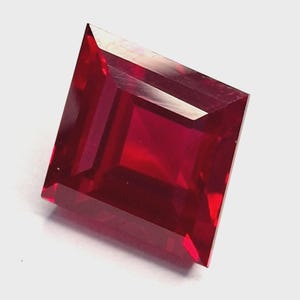 Op de afbeelding: Een gefacetteerde, vierkant geslepen robijn edelsteen. De edelsteen heeft een diepe, verzadigde rode kleur met scherpe, gedefinieerde randen en facetten die licht reflecteren. De edelsteen staat tegen een effen witte achtergrond.