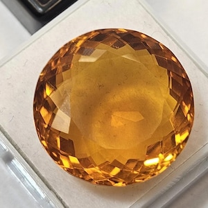 以下が含まれることがあります： 温かみのある黄金色の大きな、ファセット加工された丸い宝石。 宝石は白いプラスチックホルダーにセットされており、ファセットは光と影の鮮やかな表示を作成します。 宝石はシトリンのようです。