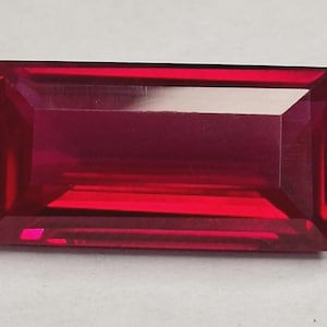 Könnte beinhalten: Ein rechteckiger, facettierter Edelstein in tiefroter Farbe. Der Edelstein hat eine polierte Oberfläche mit scharfen Kanten und eine reflektierende Qualität, was auf einen Rubin hindeutet. Der Schliff ist eine Baguetteform, ideal für Schmuck.