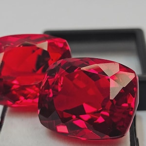 May include: Two faceted, cushion-cut, deep red gemstones. The gems are highly polished, reflecting light with brilliant facets. The gems are displayed on a white surface with a black tray in the background. These gems are suitable for jewelry making.