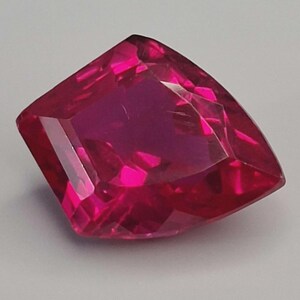 May include: A faceted, cushion-cut gemstone in a vibrant shade of ruby red. The gem's multiple facets reflect light, creating a brilliant display. The gemstone is set against a neutral background, highlighting its colour and clarity.