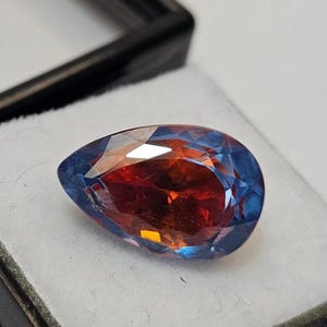 May include: A faceted, teardrop-shaped gemstone exhibiting colour change. The gem shifts from a deep red-orange in the centre to a vibrant blue around the edges. It is presented in a black display case with a white interior, highlighting its brilliance.